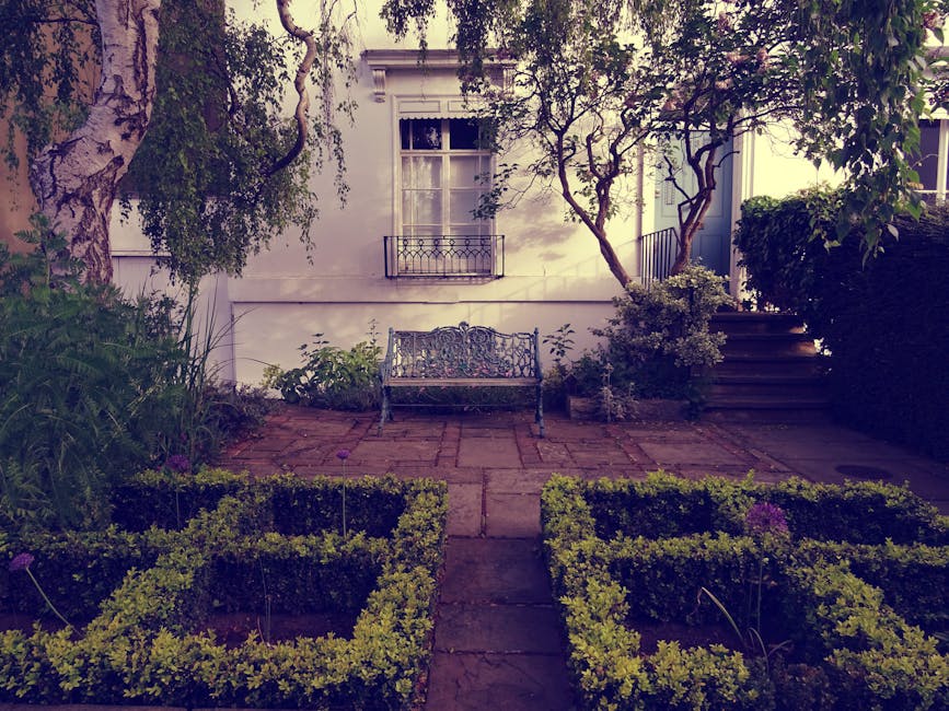 A small front garden adjacent to a white residential building featuring a paved area with weathered brick walkways. In front of the house, a decorative metal garden bench with intricate patterns is po