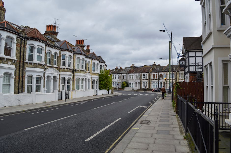 A quiet residential street in Plaistow Road West Ham featuring a row of Victorian-style terraced houses with white and brick facades, and bay windows. The foreground shows a neat, paved sidewalk borde